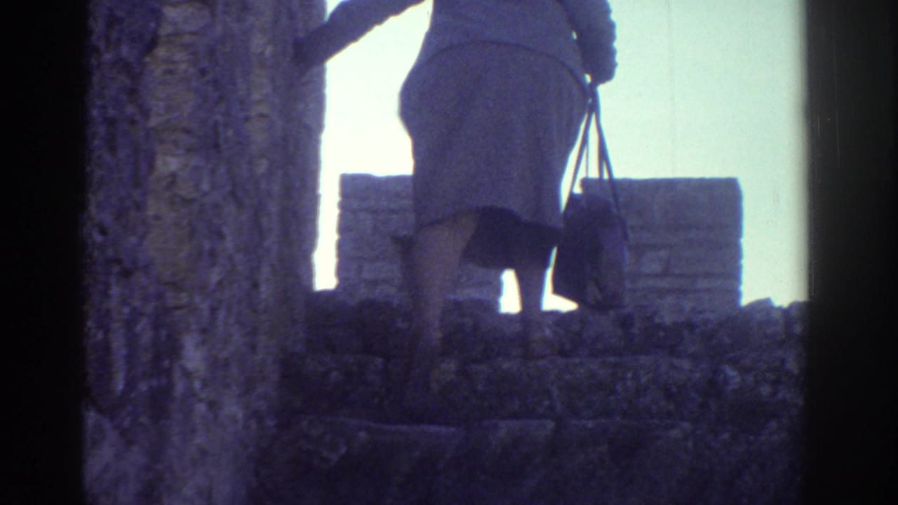In Greece home-movie footage from 1976, a woman in a light coat and hat climbs weathered stone steps while carrying a purse.