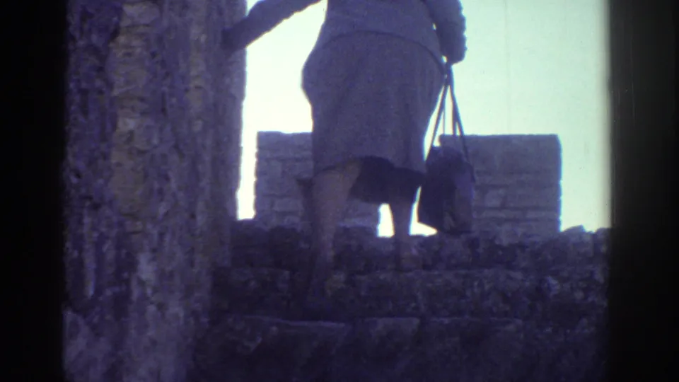 In Greece home-movie footage from 1976, a woman in a light coat and hat climbs weathered stone steps while carrying a purse.