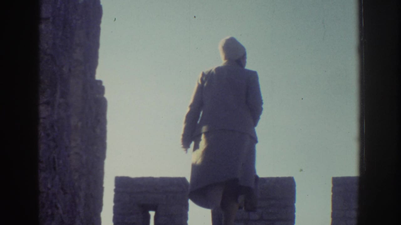 The woman appears on the upper castle wall in Greece, her coat and hat silhouetted against the bright sky.