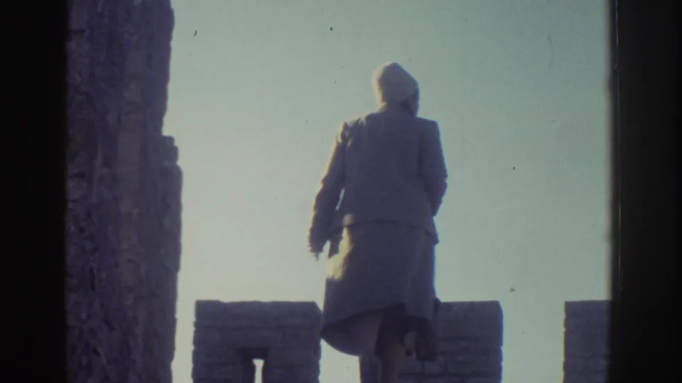 The woman appears on the upper castle wall in Greece, her coat and hat silhouetted against the bright sky.