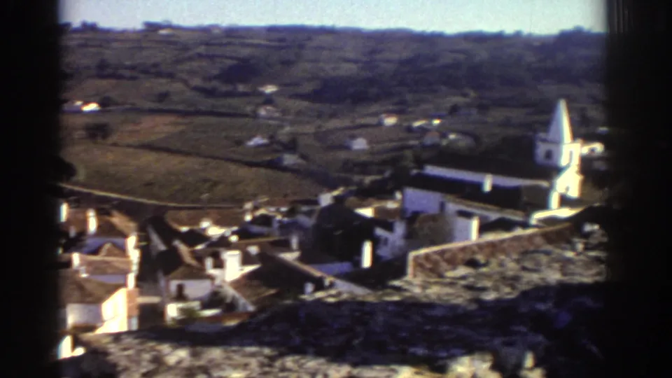 A hillside village with rooftops and a white church tower appears beyond the castle wall in Greece.
