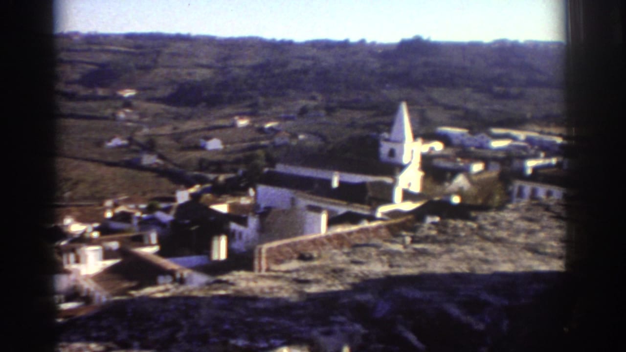 1976 Greece home-movie frame showing a hillside village overlook with white buildings, a church steeple, tiled roofs, and distant hills.