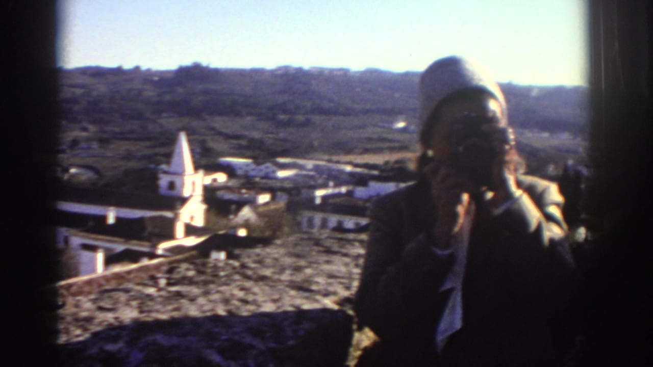1976 Greece travel footage frame with a hat-wearing traveler photographing an elevated village view with a church steeple behind them.