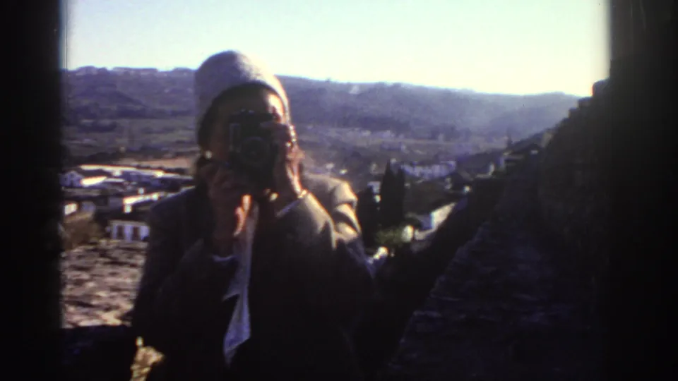 1976 Greece home-movie frame showing a traveler with a camera beside a stone overlook, with village rooftops and hills beyond.
