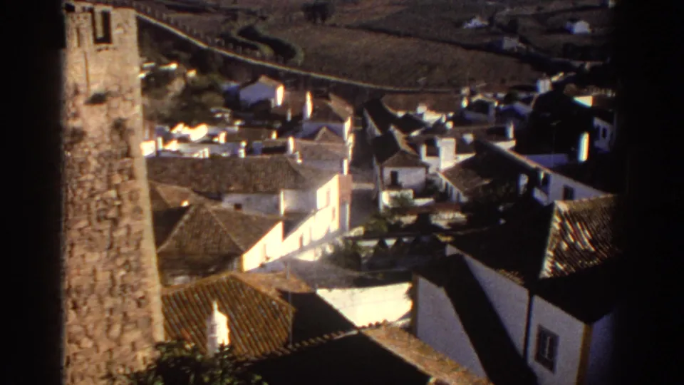 1976 Greece home-movie frame looking down over whitewashed village buildings, tiled roofs, a stone tower-like wall, and hillside terrain.