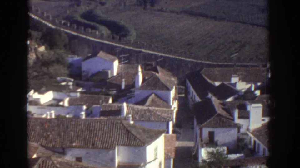 Home-movie frame of a Greek hillside town edge with tiled roofs, white walls, open fields, and a curving boundary wall.