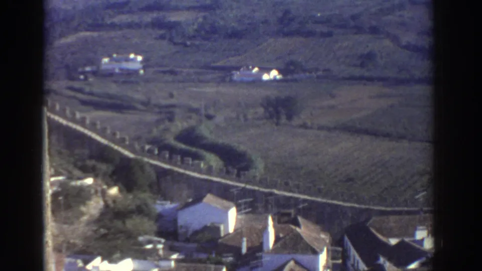 1976 Greece frame looking from town rooftops toward cultivated fields, a winding wall, and small distant rural buildings.