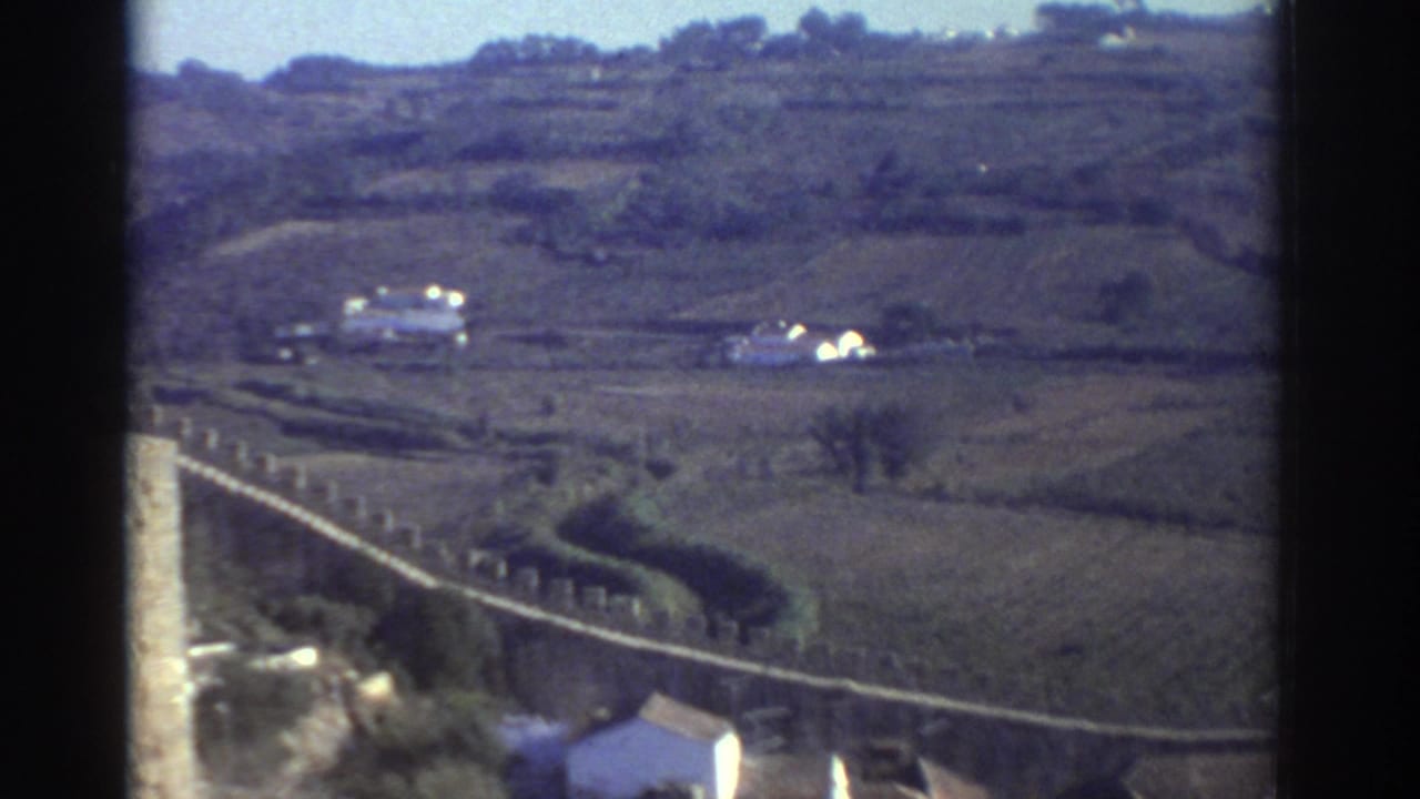 1976 Greece 8mm travel frame showing a distant town landscape with terraced hills, a pale wall or road, and small white buildings.