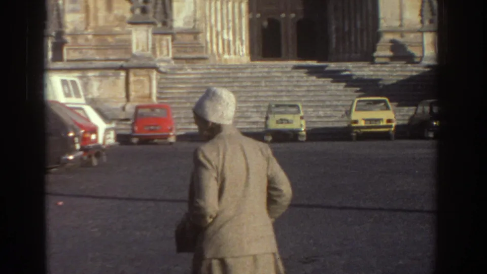 Woman in a light-colored coat and white hat crosses a Greek cathedral square toward the ornate stone entrance, with 1970s cars nearby.