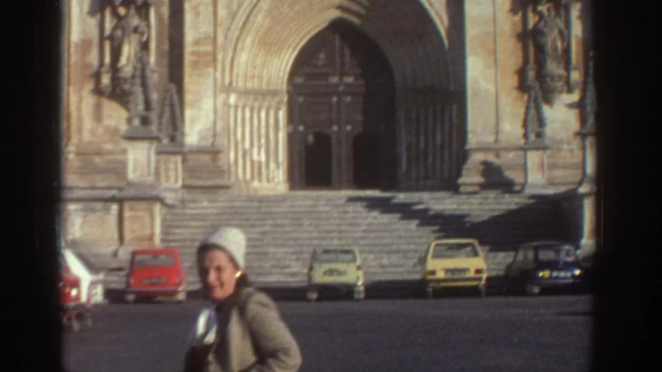 The woman is closer to camera in the cathedral square, with the broad steps, arched doorway, and several compact cars visible behind her.
