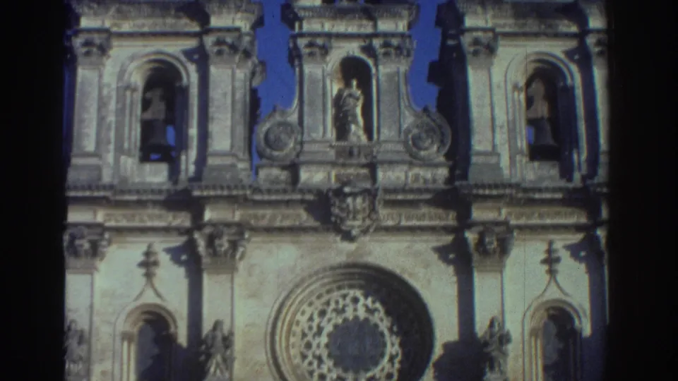 1976 Greece home-movie frame showing the upper cathedral facade with bell openings, sculptural figures, stone towers, and blue sky.