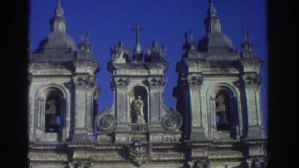 Sky-backed 1976 Greece 8mm frame of a cathedral roofline with a central cross, tower tops, bell openings, and carved statues.