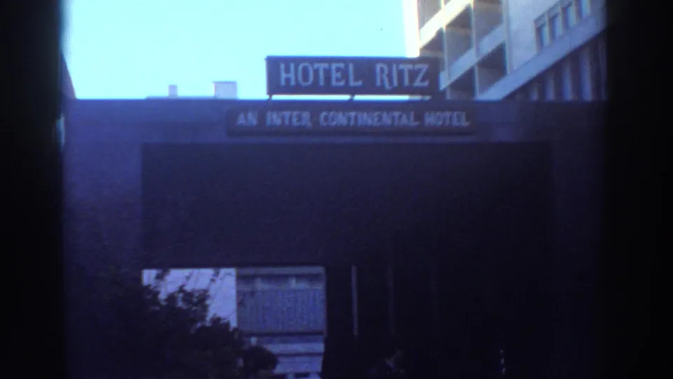 A brief urban exterior shows a Hotel Ritz sign on a building, with nearby windows and dark foreground framing.