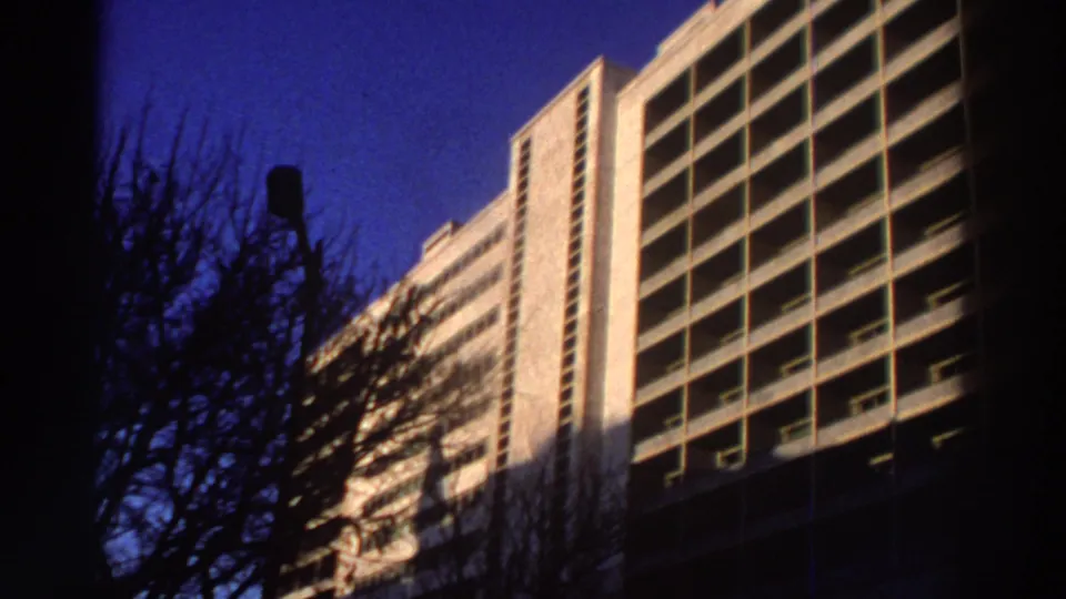 1976 Greece 8mm frame showing the hotel's modernist balcony facade under deep blue sky, framed by tree branches and a streetlamp.