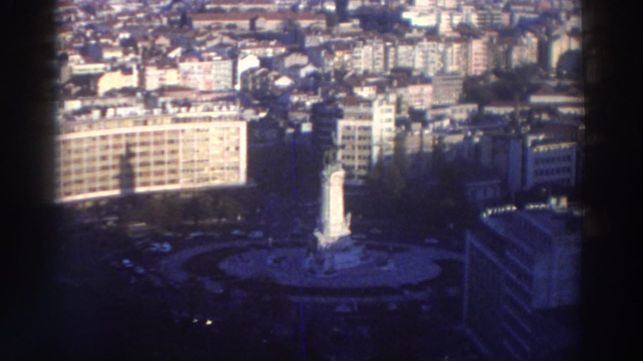 1976 8mm travel-film view over a Greek city, showing a white monument in a traffic circle, surrounding roads, and apartment blocks.