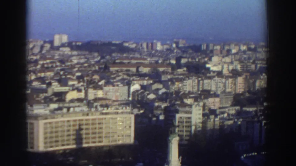 1976 8mm frame from Greece showing dense urban housing, mid-rise buildings, and part of the roundabout monument at the bottom.
