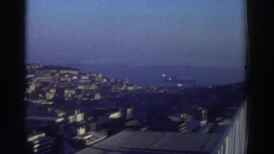 1976 Greece travel-film frame looking from the city toward a distant coastline and blue sea, with buildings in the foreground.