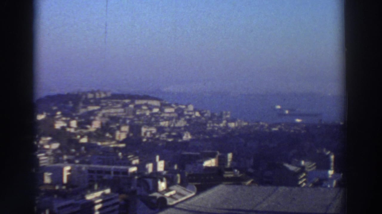 1976 Greece 8mm frame showing hillside apartment blocks, rooftops, coastline, and hazy water from a high vantage point.