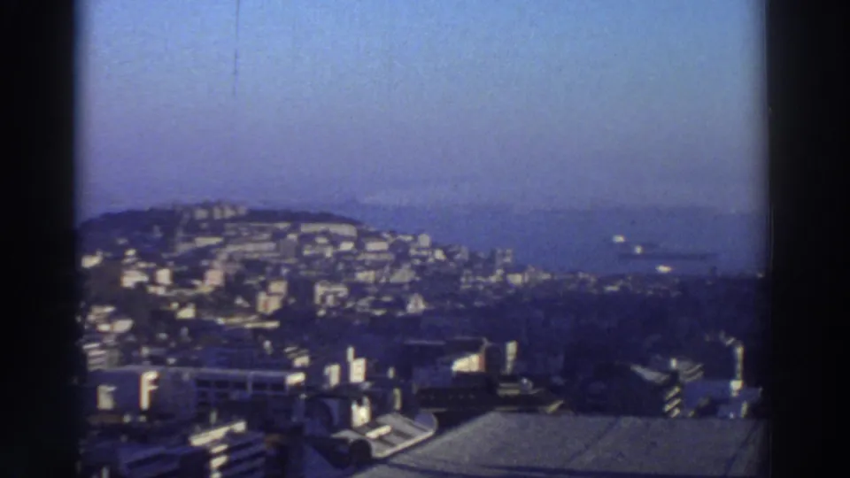 1976 Greece 8mm frame showing hillside apartment blocks, rooftops, coastline, and hazy water from a high vantage point.