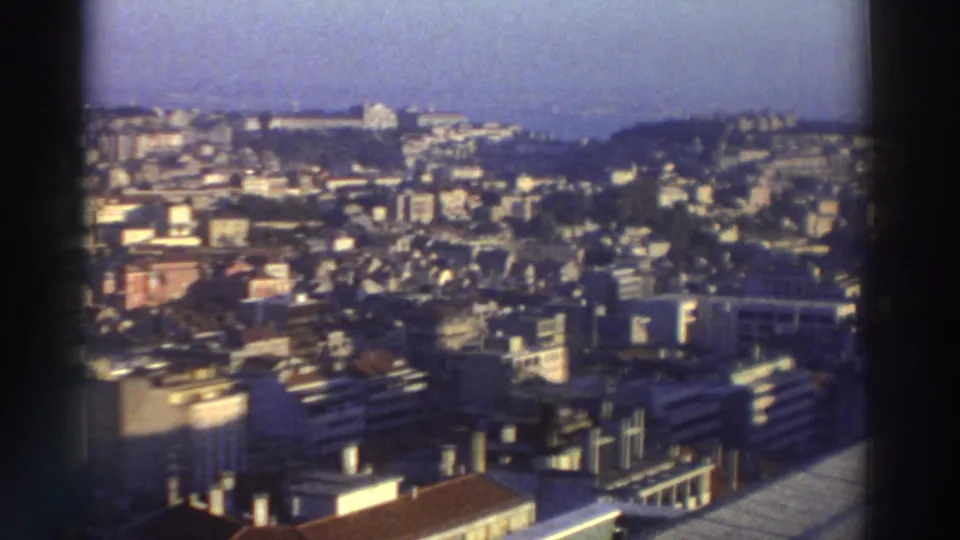 1976 Greece frame showing clustered city buildings, hillside neighborhoods, rooftops, and coastline in soft 8mm color.