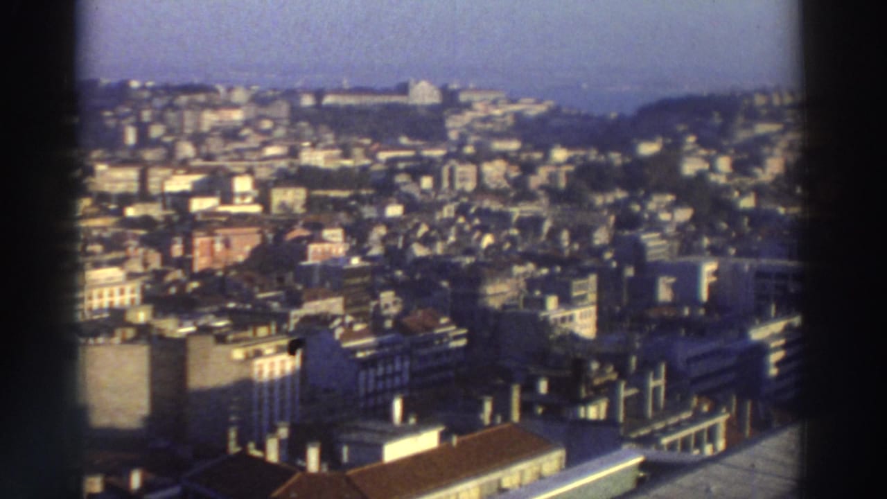 1976 Greece 8mm frame showing a high vantage view over clustered city buildings, rooftops, and distant hillside development.