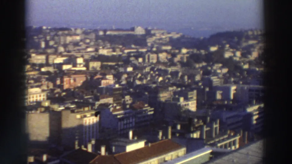 1976 Greece 8mm frame showing a high vantage view over clustered city buildings, rooftops, and distant hillside development.