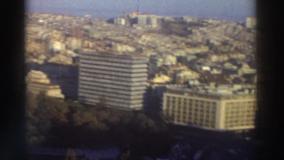 1976 Greece 8mm view of a dense urban area with two prominent modern buildings, trees in the foreground, and hillside housing behind.