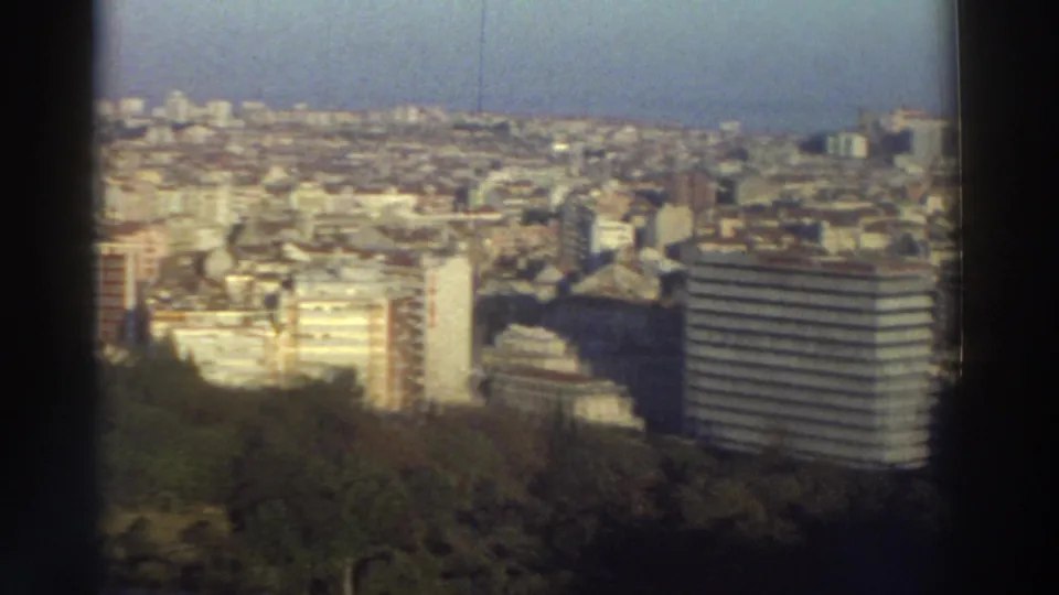 1976 Greece city panorama showing a tree-covered foreground, mixed mid-rise buildings, and a striped modern high-rise to the right.