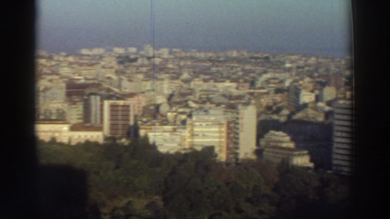 1976 Greek home-movie view across a dense city district, with trees in the foreground and dark borders suggesting filming through an opening.