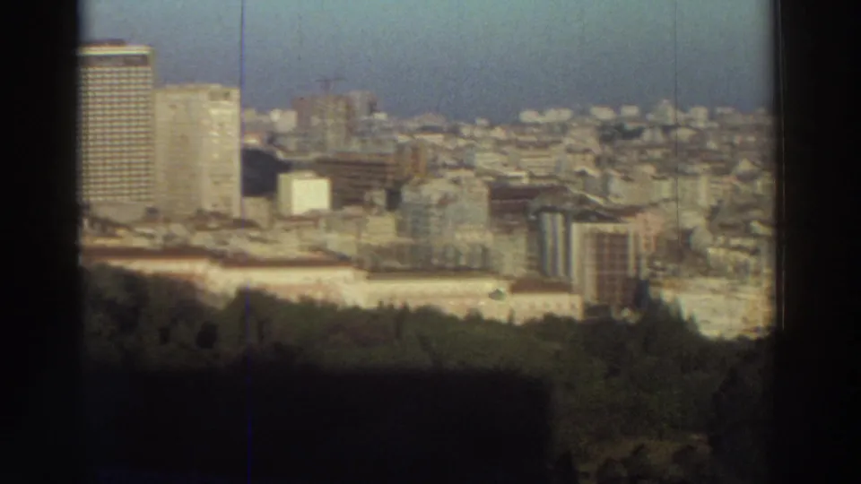 Elevated 1976 Greece view showing tall modern towers among lower city buildings, with a wooded foreground and dark framing at the edges.
