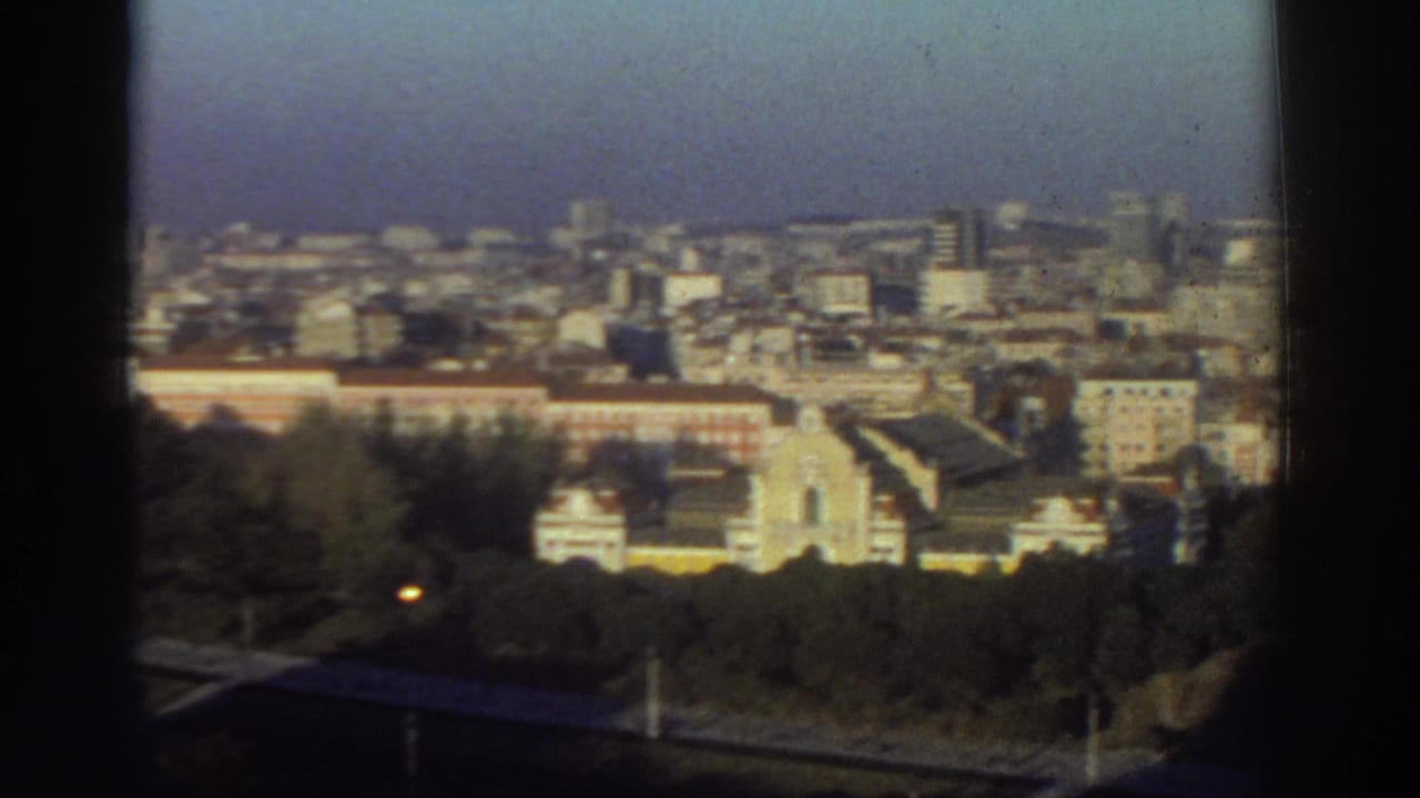 Elevated city view framed by dark window edges, with trees, pale buildings, and a hazy skyline.