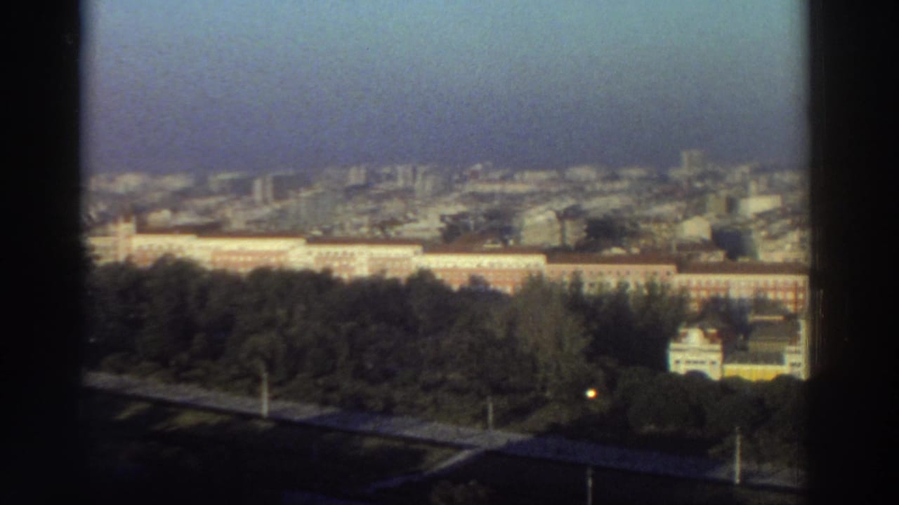 1976 Greece city panorama from a high vantage point, with trees, a long reddish building, distant high-rises, and soft film color.
