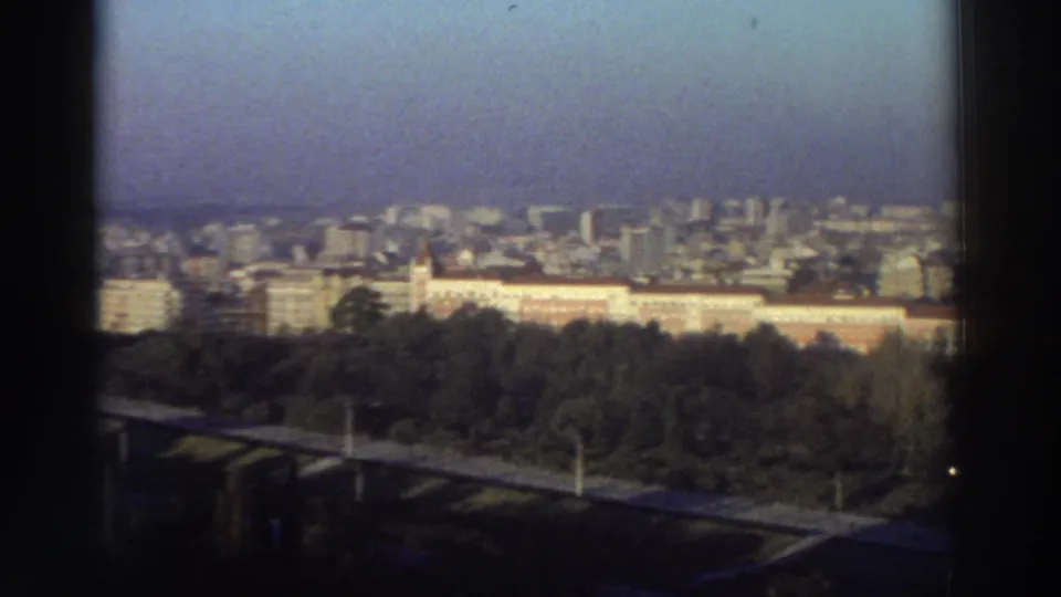1976 Greece home-movie frame showing a soft, grainy city view with foreground road infrastructure, trees, and distant urban buildings.