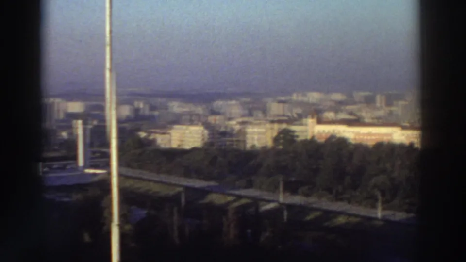 1976 Greece elevated city view with a tall foreground pole, road structure, wooded area, and distant apartment blocks in hazy light.
