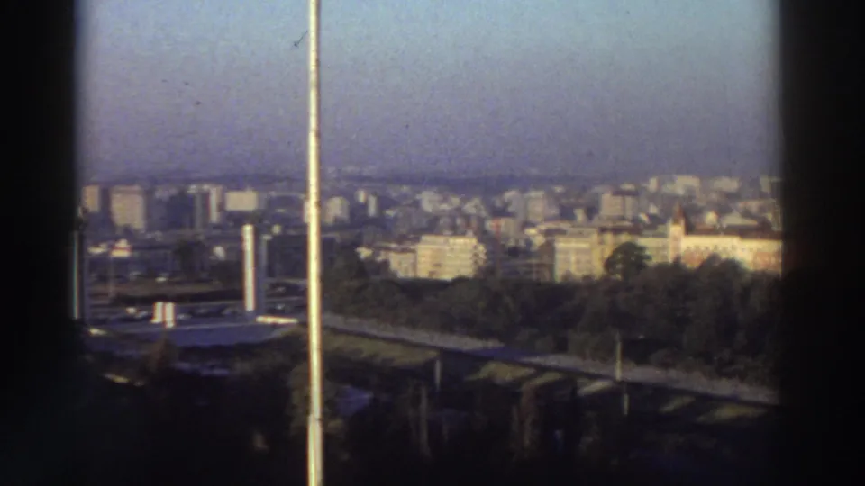 1976 8mm rooftop view in Greece showing a hazy urban skyline, trees, and pale vertical structures in the foreground.