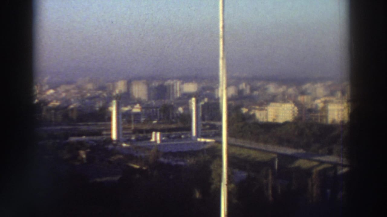 Soft 8mm view across a Greek city skyline with apartment blocks, haze, and rooftop structures in the lower foreground.