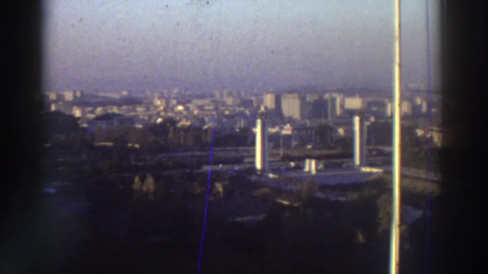 Rooftop vantage over a hazy Greek city in 1976, with white foreground structures, trees, and distant urban blocks.