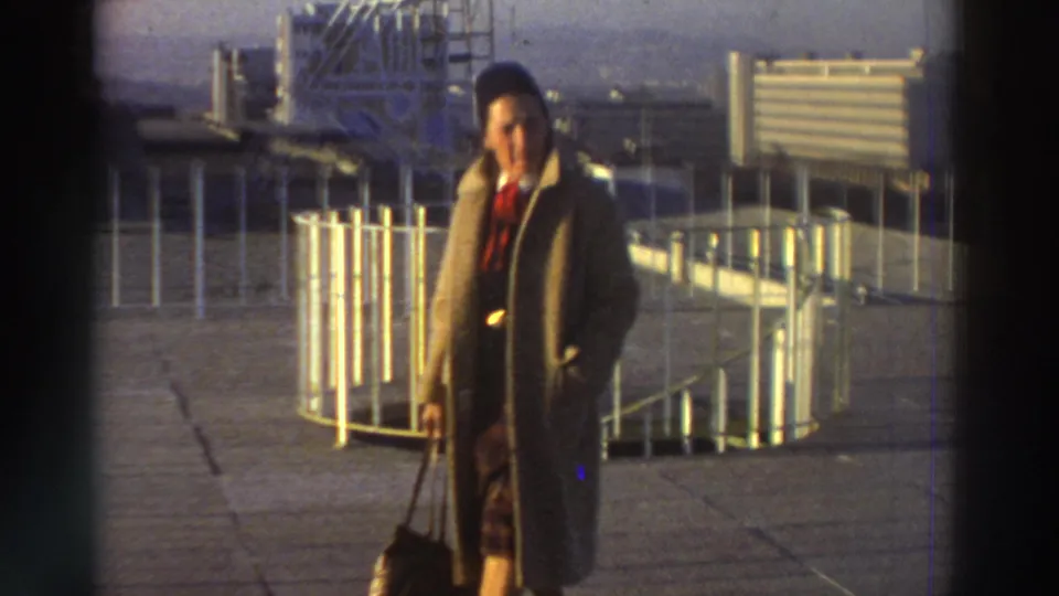 Woman in a long tan coat and dark hat stands on a rooftop in Greece, holding a bag beside white railings and industrial-looking structures.