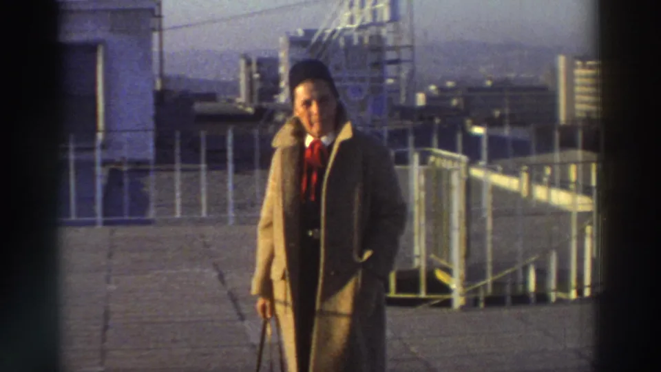 1976 Greece home-movie frame showing a man in a brown overcoat, dark hat, and red tie walking across a rooftop near railings.