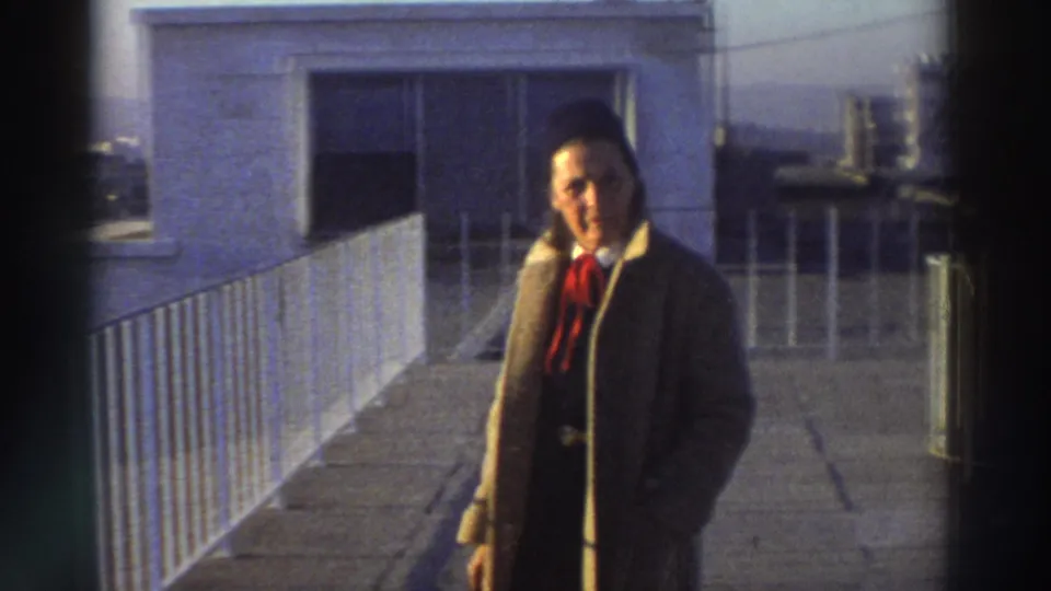 1976 Greece rooftop frame with a man in a brown overcoat walking near a white metal railing and rooftop doorway.