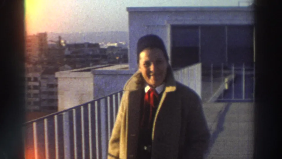 Closer 1976 Greece home-movie view of the man in an overcoat and red tie on a rooftop above city buildings.