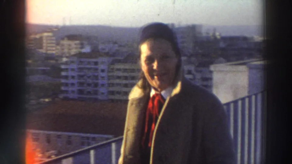 1976 Greece frame showing the man near a rooftop railing, with hazy city buildings and warm film flare at the edge.