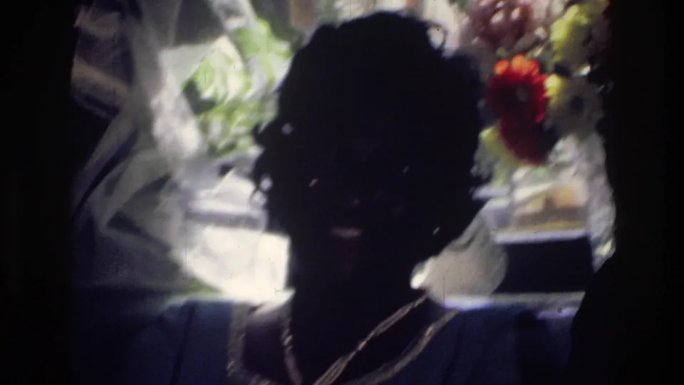 Backlit 1976 Harlem home-movie frame of a seated woman in a dim interior with flowers and window light behind her.