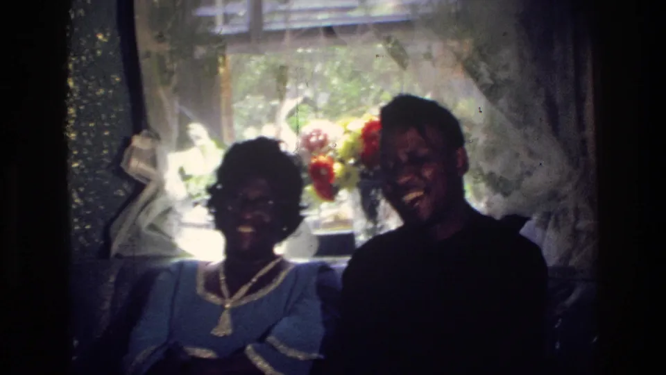 In a 1976 Harlem interior, a woman in a blue dress and a man in a dark shirt sit together smiling near a bright window.