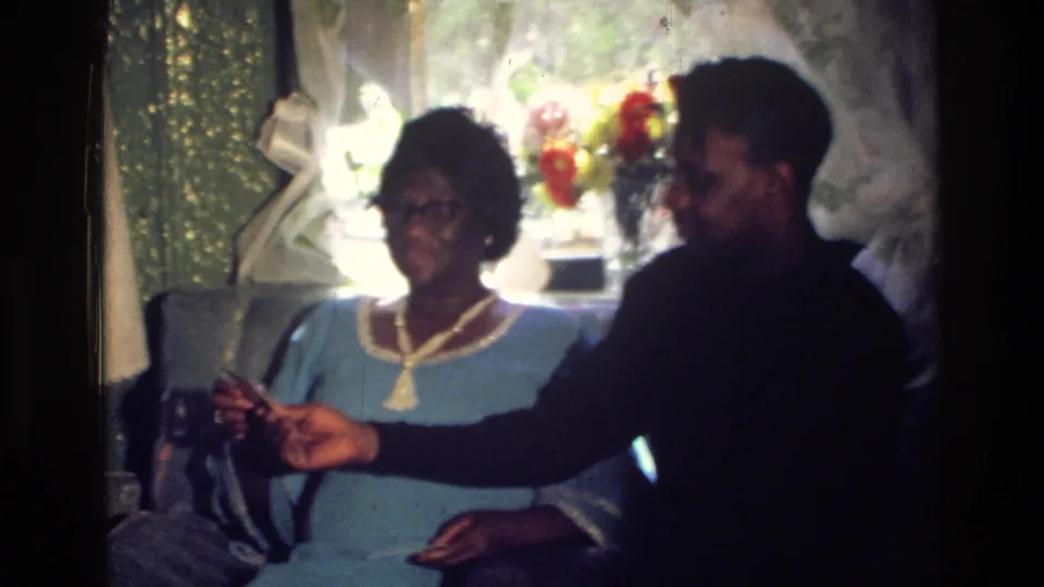 The couple sits together in a 1976 Harlem home interior as the man reaches toward a small object near the woman.