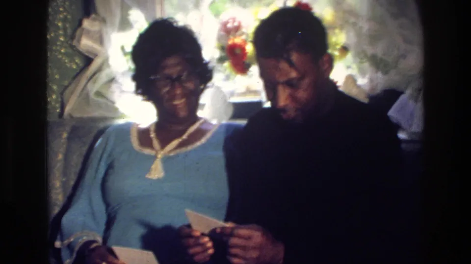 A 1976 Harlem home-movie frame shows the woman smiling while the man studies a small photograph or paper beside her.