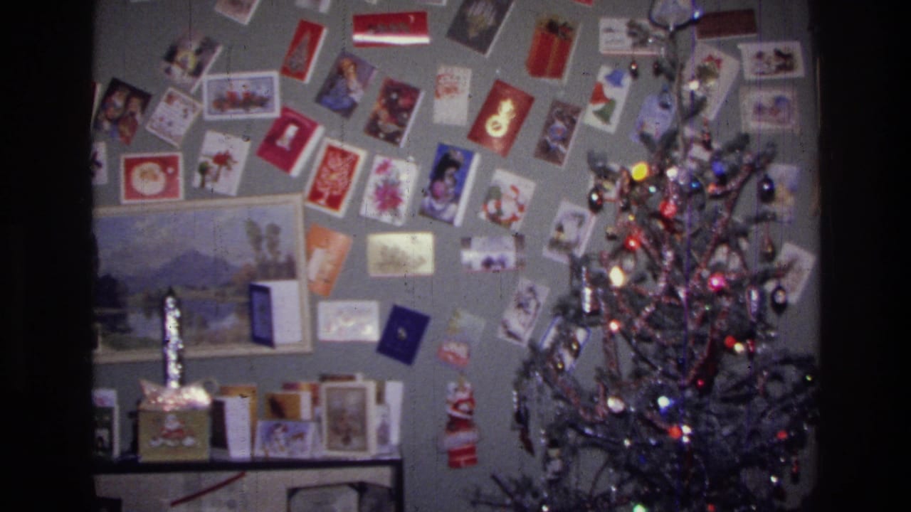 1976 Harlem home-movie frame showing a gray living room wall filled with holiday cards, a framed landscape, and a small Christmas tree.