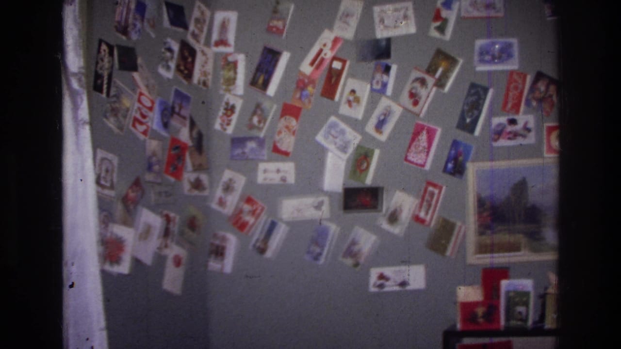 Home-movie frame from the supplied Harlem 1976 clip showing clusters of holiday cards on a gray wall with part of a framed landscape print.