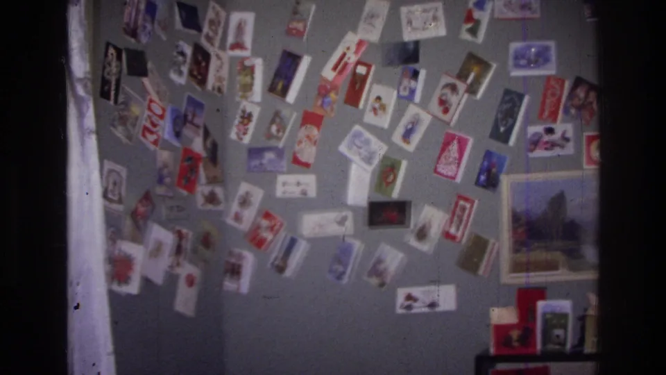 Home-movie frame from the supplied Harlem 1976 clip showing clusters of holiday cards on a gray wall with part of a framed landscape print.