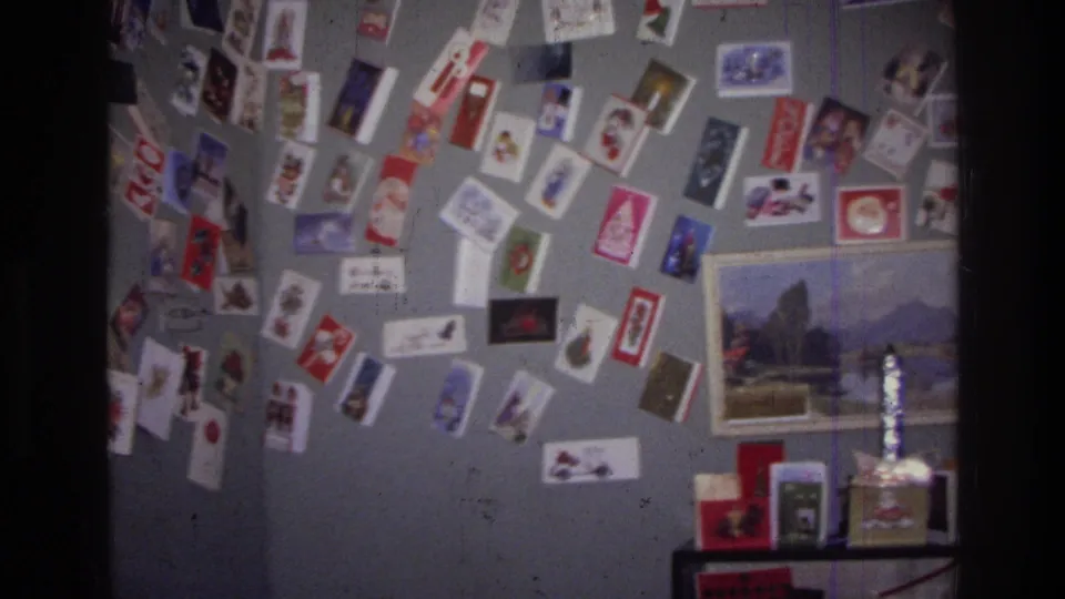1976 Harlem home-movie frame with a slightly angled view of Christmas cards, postcards, a landscape picture, and objects on a shelf below.
